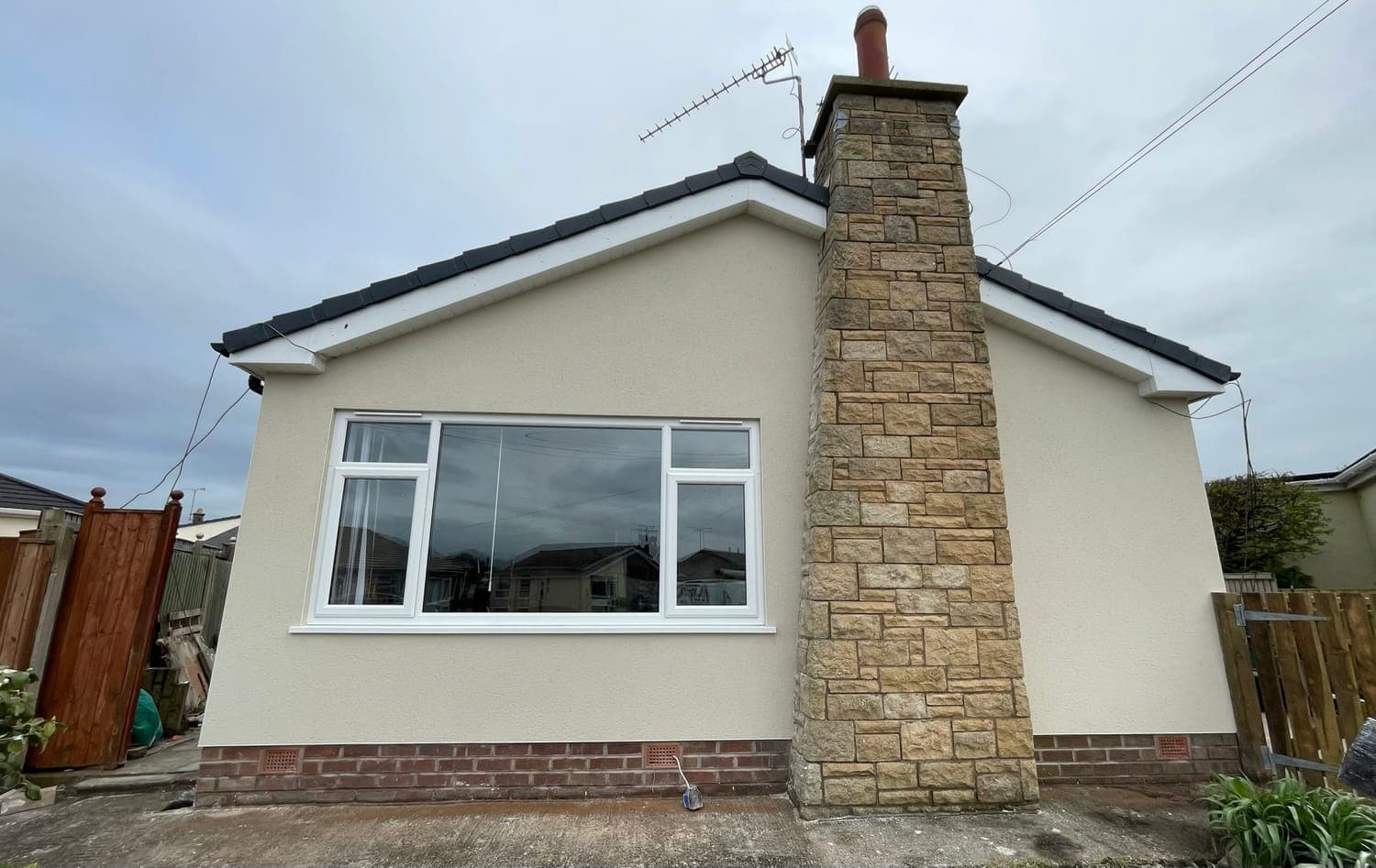 Front elevation of a bungalow in cream monocouche render, with the stone chimney and brick plinth left exposed